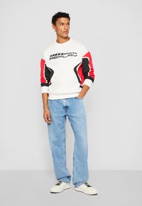 Hvit hettegenser med svarte og røde detaljer, med en stor "REPLAY"-grafikk. Kombinert med lyseblå denimjeans og hvite sneakers.