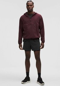 Giacca a vento con cappuccio color mogano e tasca frontale, abbinata a pantaloni corti neri. Presenta una trama leggera e liscia e una chiusura a metà zip.