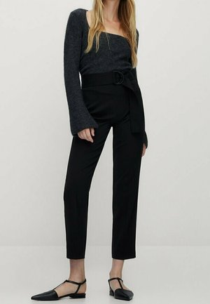 Femme portant un pantalon noir taille haute avec ceinture nouée, associé à un pull gris foncé épaules dénudées et des ballerines noires pointues.