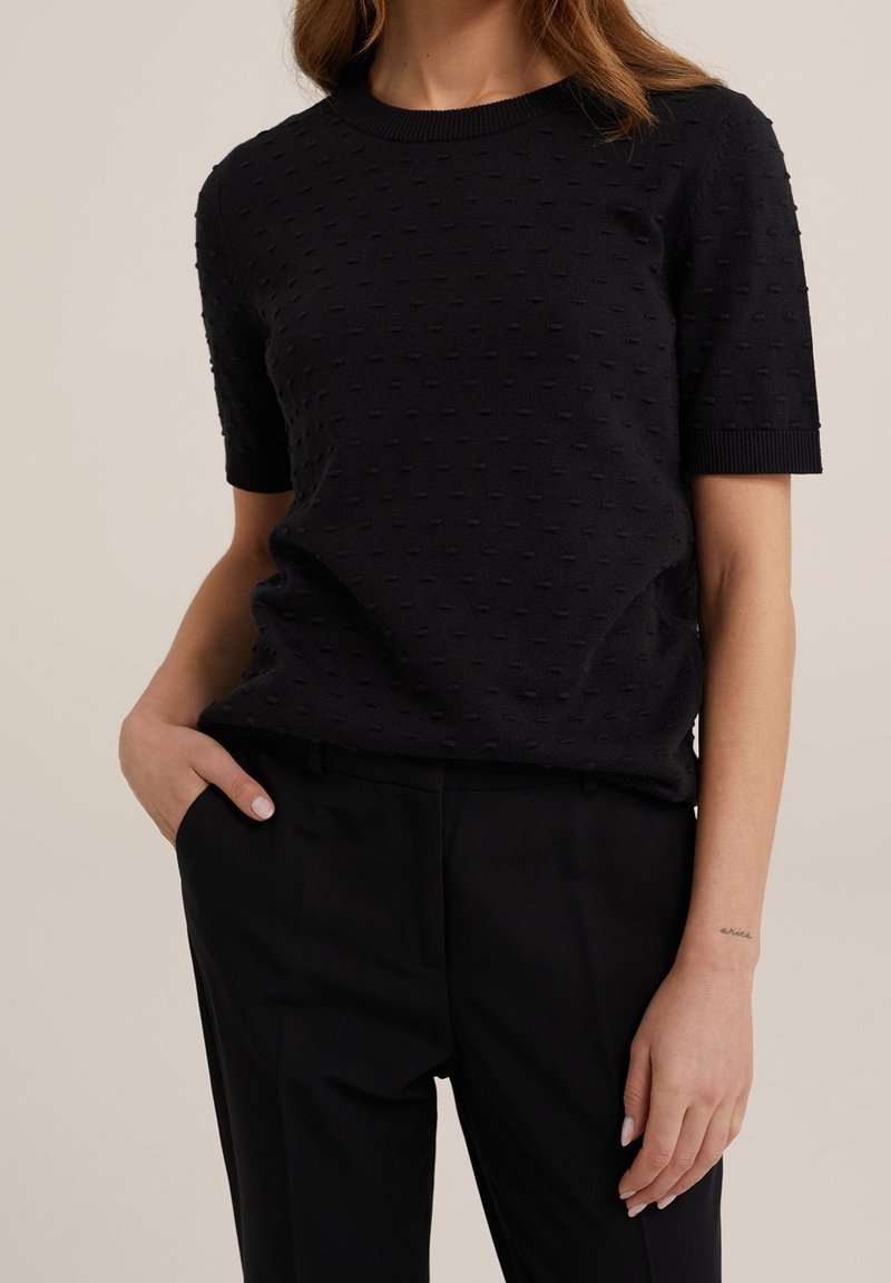 Femme portant un haut noir à manches courtes texturé et un pantalon noir, une main dans la poche et un tatouage visible sur son poignet.