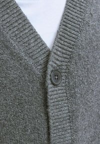 Pelēks kardigāns ar teksturētu adījumu, ar ribotu apkakli un priekšējo aizdari. Uz priekšpuses redzams viens melns plastmasas pogu.