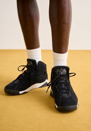 Ben iført sorte højtopede sneakers med hvide såler og hvide sokker, stående på en sennepsgul gulv.