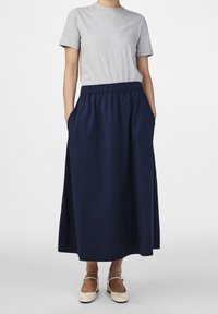 Šedé tričko s krátkým rukávem spojené s dlouhou tmavě modrou elastickou sukní s kapsami. Modelka nosí béžové boty.