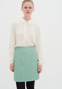 Cremefarvet bluse med knaplukning foran og puffede ærmer, parret med en grøn tekstureret nederdel. Modellen står foran en hvid baggrund.