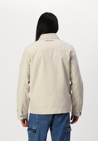 Beige cotton jacket with a pointed collar and buttoned cuffs, featuring a subtle logo printed on the back, styled with blue cargo pants.