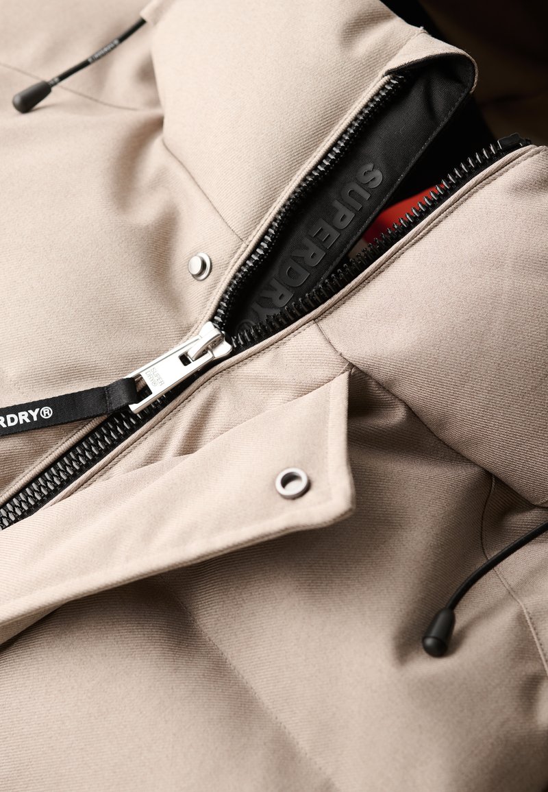 Beige jas met een ritssluiting, zilverkleurige hardware en trekkoorddetails. De stof heeft een gladde textuur met zichtbare stiksels en branding.