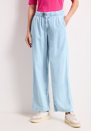 Kvinde iført lyseblå vidde jeans med elastisk talje og hvide sneakers, står med den ene hånd i lommen.