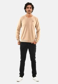 Beige Strickpullover mit V-Ausschnitt, langen Ärmeln, gerippten Bündchen und einem kleinen Logo auf der Brust, kombiniert mit schwarzen Slim-Fit-Jeans und weißen Sneakern.