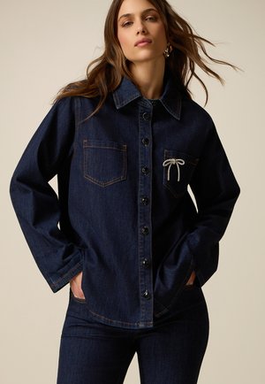 Kobieta nosząca ciemnoniebieską kurtkę jeansową z guziczkami z przodu, dwoma kieszeniami na piersiach i dekoracyjną białą kokardą na prawej kieszeni, ręce w kieszeniach.