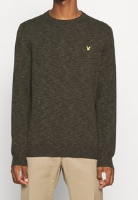 Tummanvihreä neulepusero, jossa pyöreä pääntie, resorit hihansuissa ja helmassa. Vasemmassa rinnassa pieni keltainen logo. Pehmeä tekstuuri, rento muotoilu.