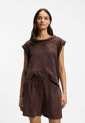 Frau mit langen dunklen Haaren, die ein ärmelloses, lockeres braunes Oberteil mit weitem Kragen und passende Shorts mit Kordelzug vor weißem Hintergrund trägt.