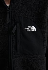 Black fleece jacket featuring a textured upper fabric, smooth pocket with a zipper, and white embroidered logo on the chest.