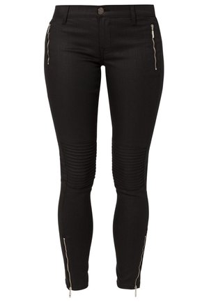 Kvinde iført en hvid tætsiddende skjorte med guld- og sorte bælte-designs, sorte skinny bukser med knædetaljer, sorte skinnende loafers og en lille crossbody taske.
