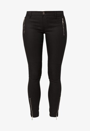 Kvinde iført en hvid tætsiddende skjorte med guld- og sorte bælte-designs, sorte skinny bukser med knædetaljer, sorte skinnende loafers og en lille crossbody taske.