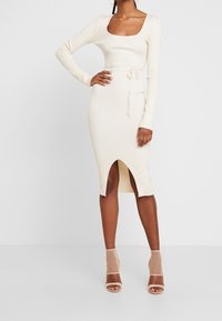 Strickkleid in Creme mit langen Ärmeln und quadratischem Ausschnitt, ausgestattet mit einer taillierten Taille, einem Bindetail und einem oberschenkelhohen Schlitz vorn. Sandalen mit transparentem Absatz.