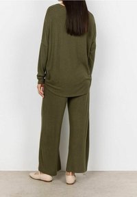 Frau steht mit dem Rücken zur Kamera, trägt ein locker sitzendes, olivgrünes Langarmoberteil und weite Hosen mit beigen Ballerinas auf einem Betonboden.