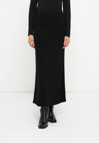 Svart, lang strikket skjørt med en tettsittende midje og en rett silhuett. Kombinert med svarte ankelstøvler. Glatt tekstur, minimalistisk design.