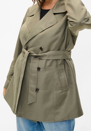 Vrouw draagt een olijfgroene trenchcoat met ceintuur en dubbele knopen over een blauwe spijkerbroek, arm geheven naar het hoofd.