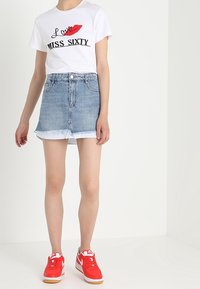 Maglietta bianca con grafica di labbra rosse e testo nero, abbinata a una gonna di jeans azzurri chiari con orlo sfilacciato e look usato, scarpe da ginnastica rosse.