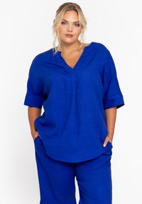 Blauw linnen top met een V-hals, korte opgerolde mouwen en een losse pasvorm. Bevat twee zijzakken en een gestructureerde stof.