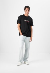 Black cotton t-shirt with red and white "tommy" logo, paired with light blue straight-leg jeans and white sneakers. Minimalist background.