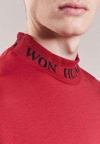 Jersey de algodón rojo con cuello alto que presenta un diseño acanalado en el cuello y texto negro. Textura suave, diseño ajustado y color sólido sin patrones ni acentos.