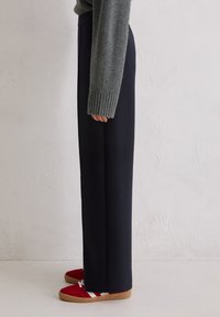 Granatowe spodnie o szerokich nogawkach z gładką fakturą, w połączeniu z czerwonymi trampkami z białymi paskami i gumową podeszwą, widok z boku.