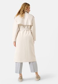 Manteau beige avec taille ceinturée, design à double boutonnage et détail à l'arrière en couches. Porté sur un pantalon large gris clair.