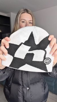 Pleteni čepici v beli in črni barvi z vzorcem zvezd in logotipom "HUF"; ima mehko teksturo in okroglo obliko.