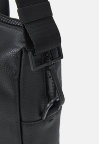 Sac en cuir noir texturé avec fermeture éclair, doté d'une sangle durable et d'accents en métal noir. La vue de côté met en évidence le détail de la fermeture éclair.