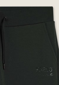 Pantalons de survêtement vert foncé avec une taille côtelée, un cordon de serrage et un texte décoratif en strass sur le bas à l'avant.