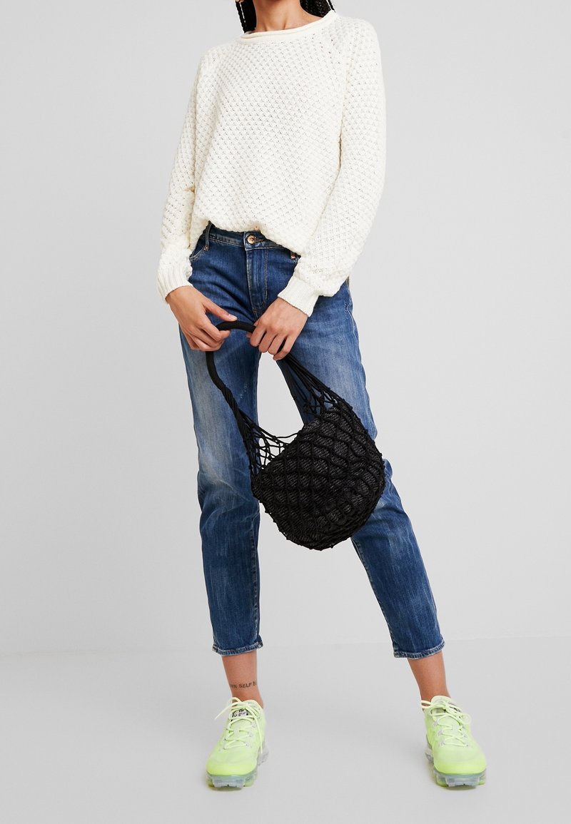Personne portant un pull texturé blanc, un jean court bleu, des baskets vert néon, tenant un sac à main noir en filet avec les deux mains.