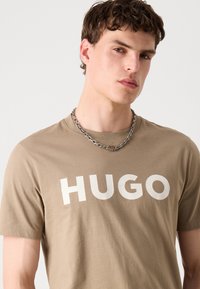 Bežové bavlněné tričko s krátkým rukávem, na přední straně s velkým bílým nápisem "HUGO". Viditelný výrazný stříbrný řetízek.