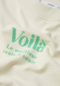T-shirt en coton couleur crème avec encolure côtelée. Présente un texte vert : "Voilà Le meilleur testé à venir" affiché de manière bien visible. Texture douce.