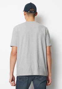 Tricou gri, cu mânecă scurtă și croială lejeră. Textură netedă din bumbac, cusături minimale, fără branding vizibil. Purtați cu blugi închiși și o șapcă.