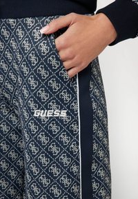 Jūras spēku raksta sporta bikses ar visā virsmā redzamu logo dizainu, sānu kabatu un balto izšūto "GUESS" logo priekšpusē.