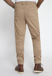 Khaki broeken met een taps toelopende pasvorm, een elastische tailleband en twee achterzakken met gestikte details. G gedragen met bruine leren laarzen.