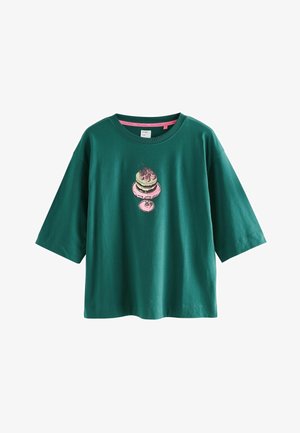 Vihreä puuvillapaita lyhyillä hihoilla, jossa on graafinen kuva moniportaisesta kakusta, jonka päällä on kirsikoita vaaleanpunaisena ja valkoisena.