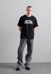 T-shirt nera con logo bianco "GCDS", abbinata a pantaloni larghi grigi. Il modello è in piedi su una superficie grigia contro un muro chiaro e semplice.