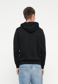 Felpa nera con cappuccio e cordini, realizzata in tessuto morbido. Maniche e orlo aderenti, mostrata da dietro con jeans azzurri chiari.