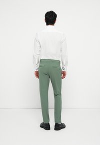 Green trousers with a slim fit, back pockets, and a smooth texture, paired with a white long-sleeve shirt and black shoes.
