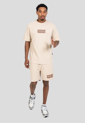 Hombre con camiseta beige "Wrong Friends" y pantalones cortos, con zapatillas blancas y negras, caminando hacia adelante sobre un fondo gris.