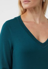 Kobieta w turkusowym swetrze z dekoltem w serek i subtelnym fakturowanym wykończeniem, widoczna od ramion do brody, na jednolitym jasnym tle.