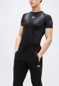 Μαύρο αθλητικό πουκάμισο με υφασμένους σχεδιασμούς και κοντά μανίκια, σε συνδυασμό με μαύρα τζόκερ με κορδόνι στη μέση και χρυσές λεπτομέρειες λογότυπου.