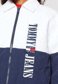 Navy and white windbreaker with a zip front, featuring "TOMMY JEANS" embroidered in blue and a branded label in red and white.