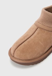 Chausson en suede marron avec un col en tricot côtelé, présentant un bout arrondi et un détail de couture visible sur le dessus et la semelle.