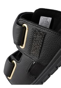 Svarta lädersandaler med två breda remmar med guldfärgad detalj, texturerad yta och gummisulor, vilket understryker hållbarhet och komfort.