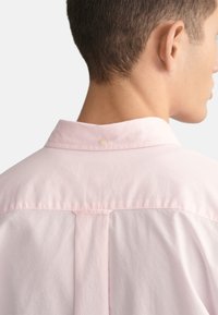 Helles rosa Button-Up-Shirt mit weicher Textur. Es verfügt über einen klassischen Kragen, dezente Faltendetails am Rücken und einen einzelnen Knopfverschluss hinten.