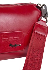 Rote Lederhandtasche von Pepe Jeans mit geprägtem Markenlabel, Metall-Logo und breitem verstellbarem Stoffriemen mit "PEPE JEANS"-Text.