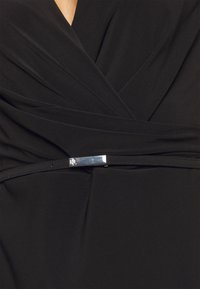 Černé šaty s plisovanou látkou, hlubokým výstřihem do V a stříbrným kovovým detailem v pase. Hladká textura po celém povrchu.